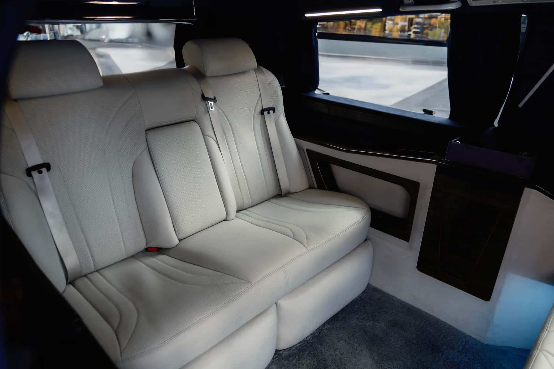 The rear bench of a people carrier in cream perforated leather, with a dark veneered cabinet and a privacy curtain at the window.